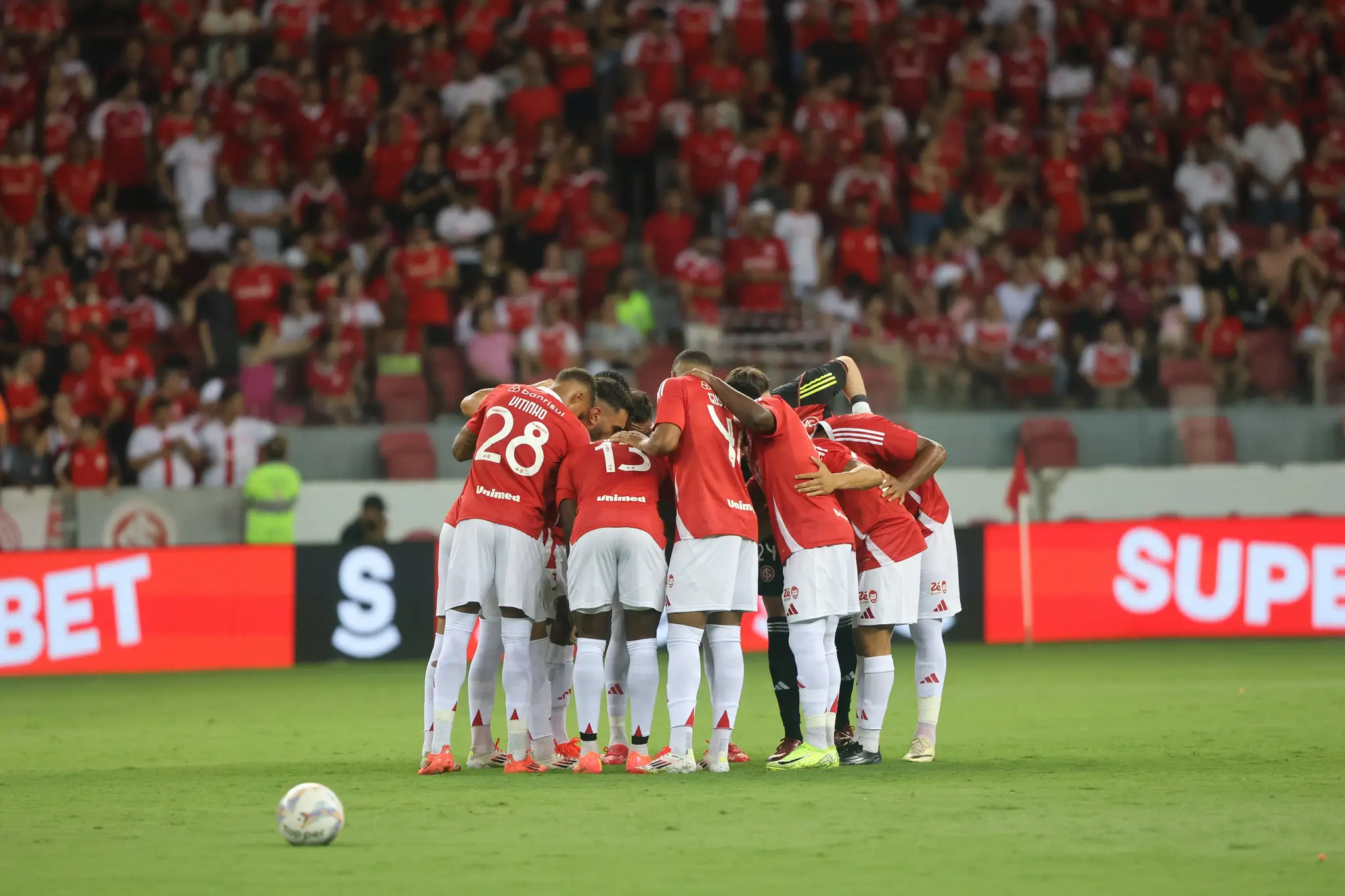 O grupo de jogadores do Internacional, vestidos com o tradicional uniforme vermelho de jogo, se abraçam em campo, no estádio Beira Rio.