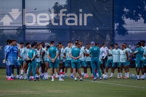 Grupo de jogadores e equipe técnica do Cruzeiro Futebol Clube, reunidos em círculo no gramado do campo de treinamento na Toca da Raposa, durante pau